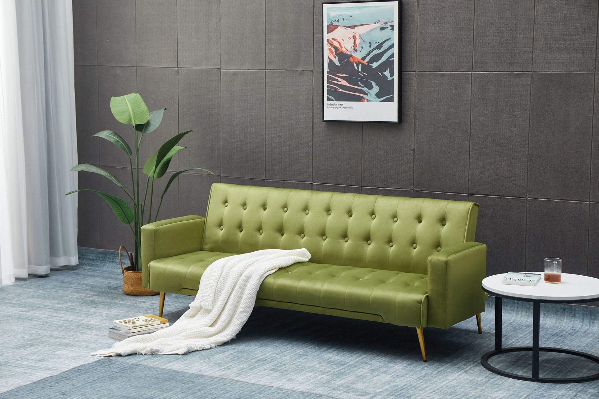 A modern living room featuring the Comfort Compact Design Sofa Bed in green, topped with a white blanket, next to a potted plant, a round side table holding a book and glass, and abstract artwork on a gray textured wall.