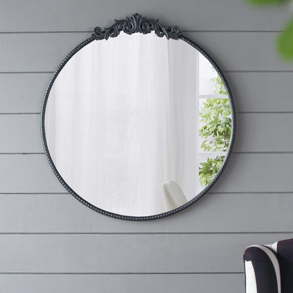 A Classic Design Mirror with a Baroque inspired frame hangs on a gray shiplap wall, reflecting a bright window with sheer white curtains and green plants. A black-and-white chair is partly visible in the lower right corner.