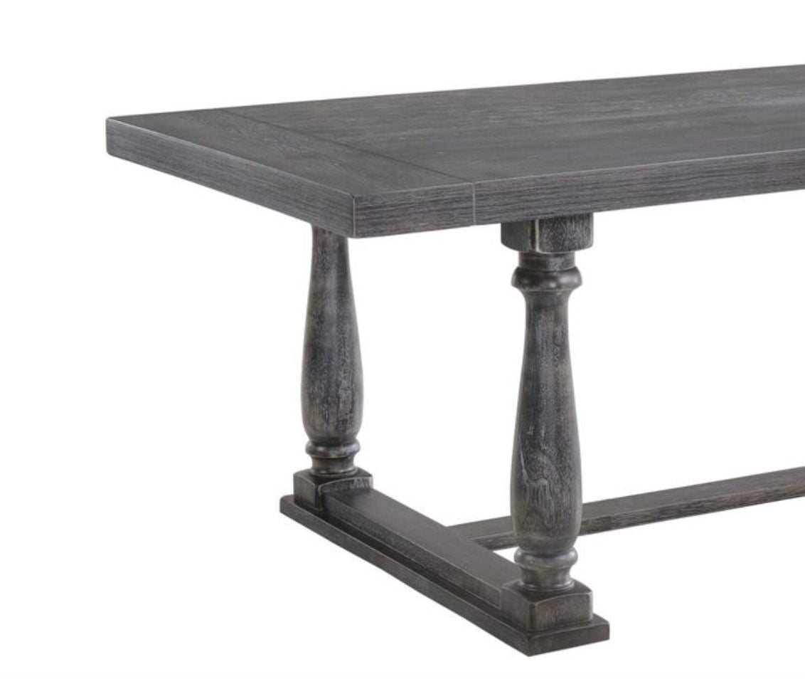 The Bernard - Weathered Dining Table features a dark wooden rectangular top and two turned pedestal legs connected by a horizontal base.