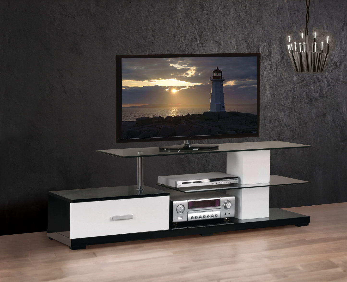 The Ivana - TV Stand - White / Black features a flat-screen TV showing a sunset lighthouse, two glass shelves for electronics, and a drawer, set against a dark textured wall with chandelier lighting and wooden flooring.