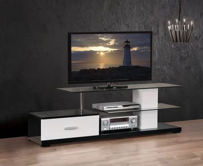 The Ivana - TV Stand - White / Black features a flat-screen TV showing a sunset lighthouse, two glass shelves for electronics, and a drawer, set against a dark textured wall with chandelier lighting and wooden flooring.