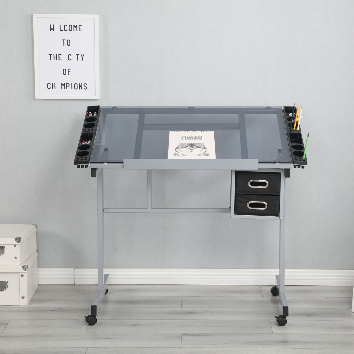An Adjustable Art Drawing Desk Craft Station Drafting With 2 Non-Woven Fabric Slide Drawers And 4 Wheels - Silver holds an open sketchbook and art supplies. White storage boxes sit to its left, below a "WELCOME TO THE CITY OF CHAMPIONS" sign on the wall.
