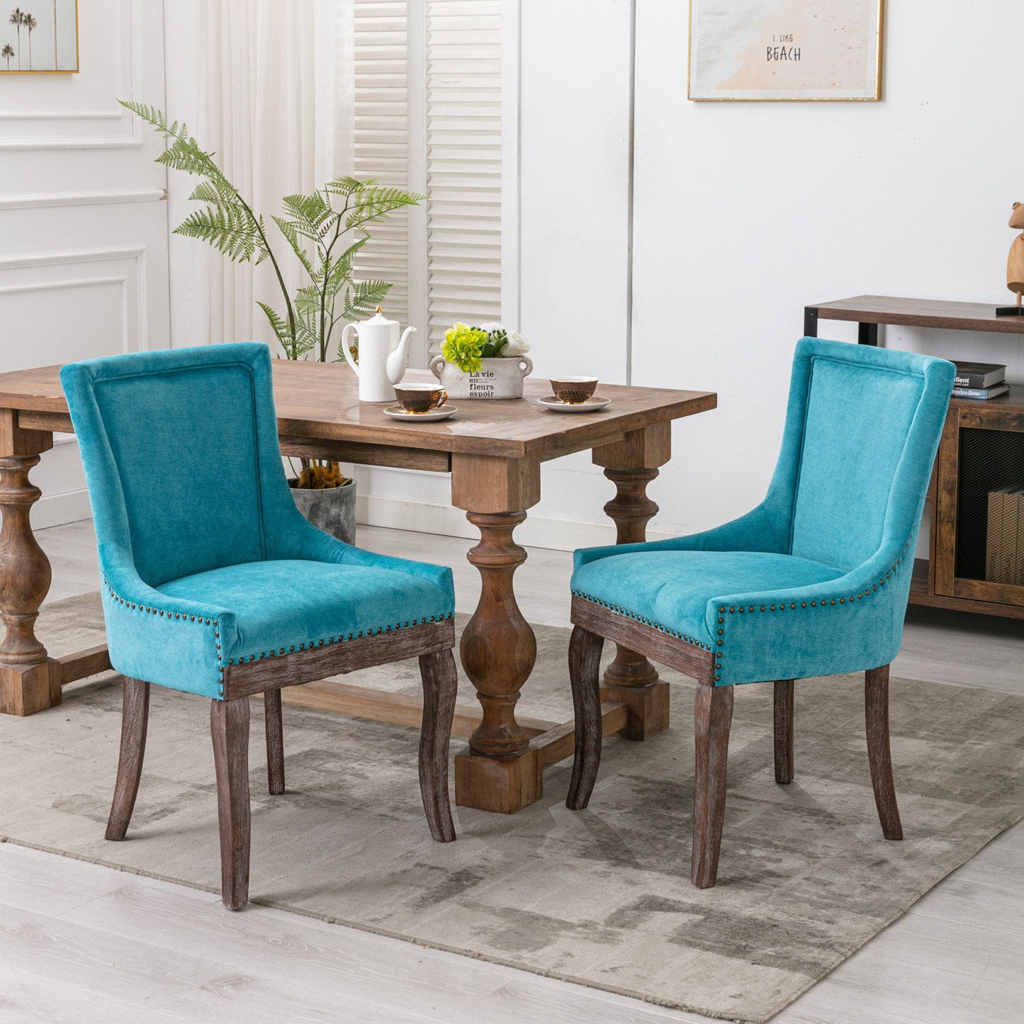 Two Ultra Side Dining Chairs with thickened teal fabric and neutrally toned solid wood legs, accented by bronze nail heads, are placed around a wooden dining table set for tea in a bright dining room with light walls and a rug.
