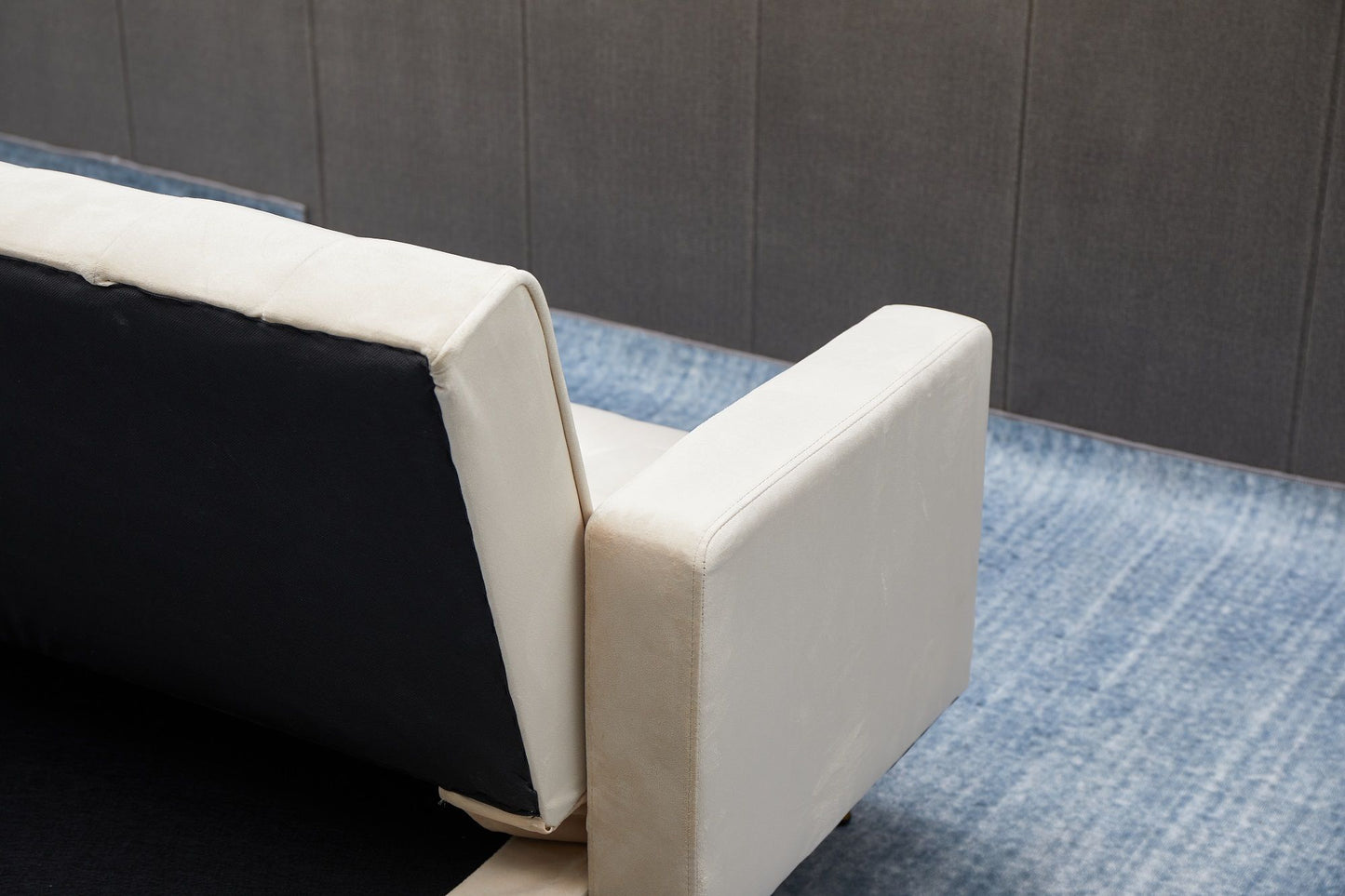 A close-up of the Comfort Compact Design Sofa Bed with white armrest and backrest, set on a blue gradient rug in front of a gray paneled wall.