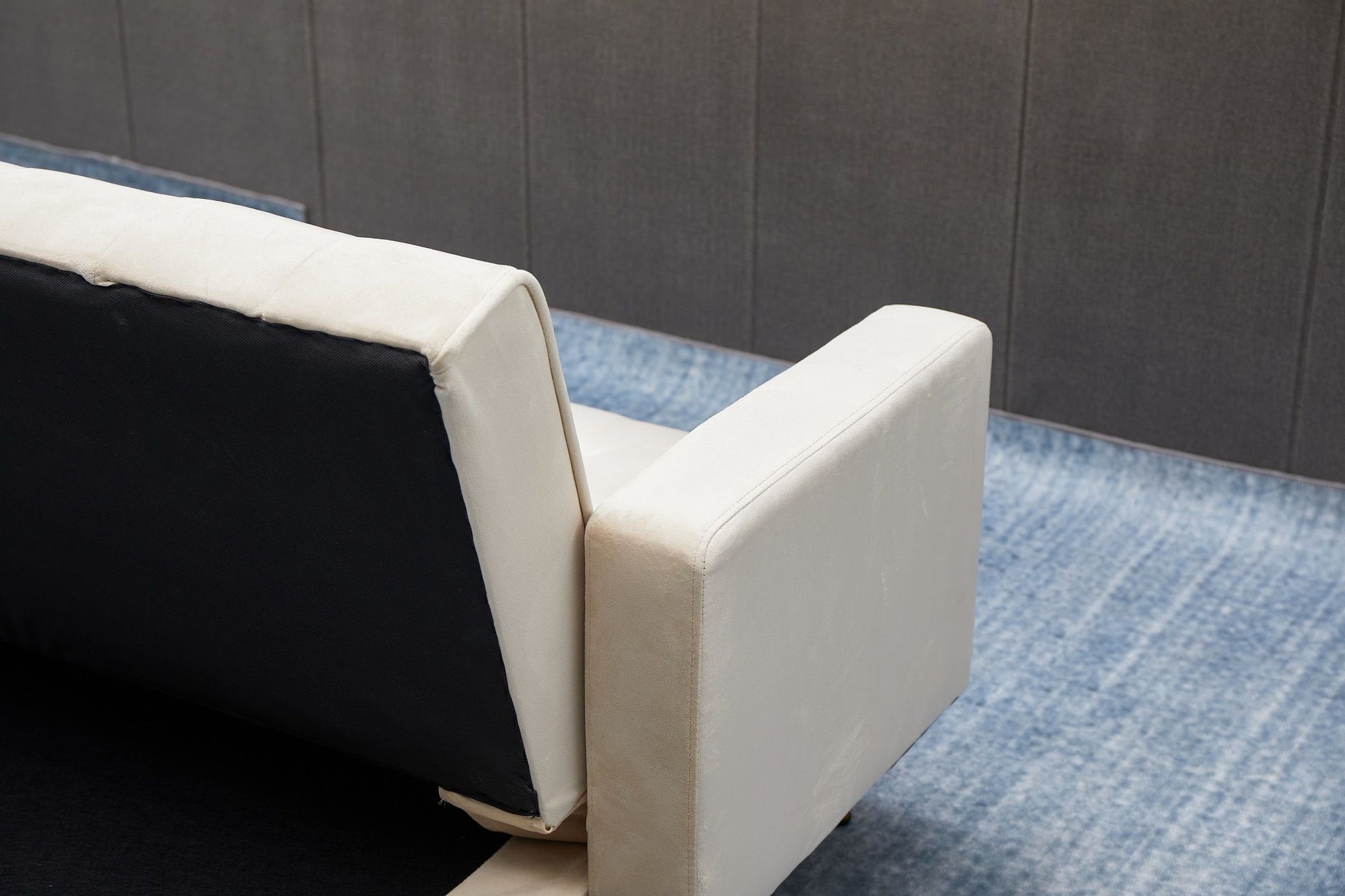 A close-up of the Comfort Compact Design Sofa Bed with white armrest and backrest, set on a blue gradient rug in front of a gray paneled wall.