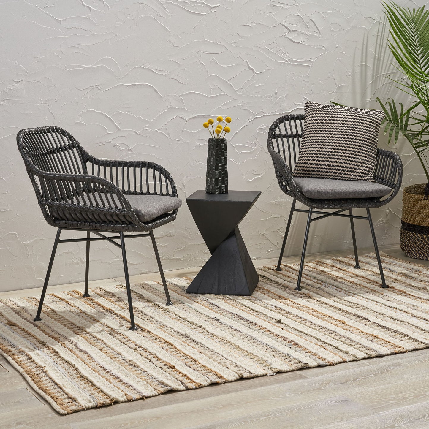 Two Outdoor Rattan Accent Chairs With Metal Legs and gray cushions sit on a striped rug, separated by a black side table with yellow flowers; a green plant and woven basket accent the textured white wall background.