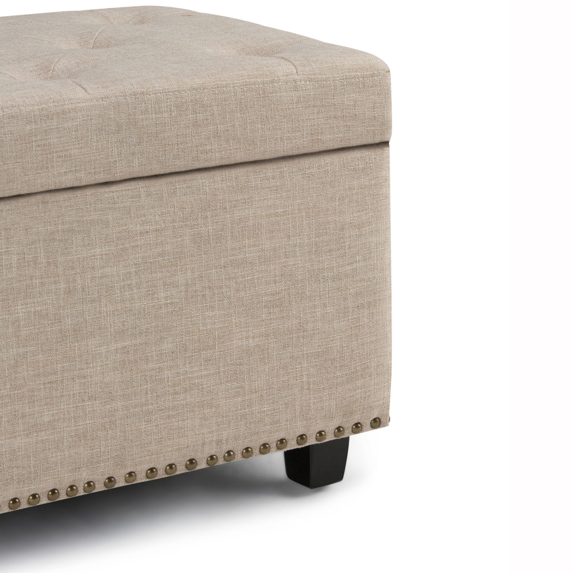 Close-up of the Hamilton Upholstered Storage Ottoman in beige, featuring a tufted top, decorative nailhead trim at the base, and black wooden legs, set against a white background.