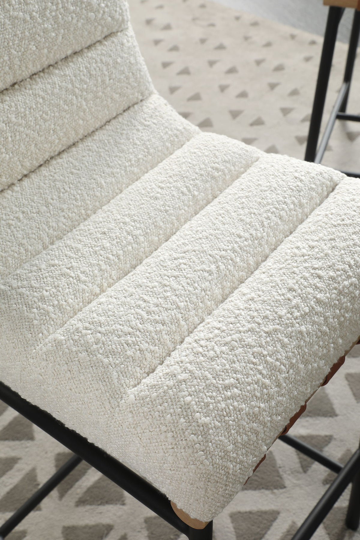 A close-up of the Lahni - 17" Boucle Fabric Counter Height Chair featuring textured off-white upholstery with horizontal tufted padding and black metal legs. A light-colored patterned rug is visible beneath the chair.