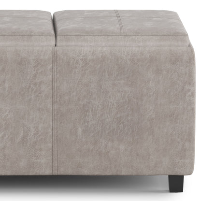 A close-up of the Avalon - Upholstered Storage Ottoman in light gray, featuring visible stitching, black legs, and a textured leather-like upholstery.