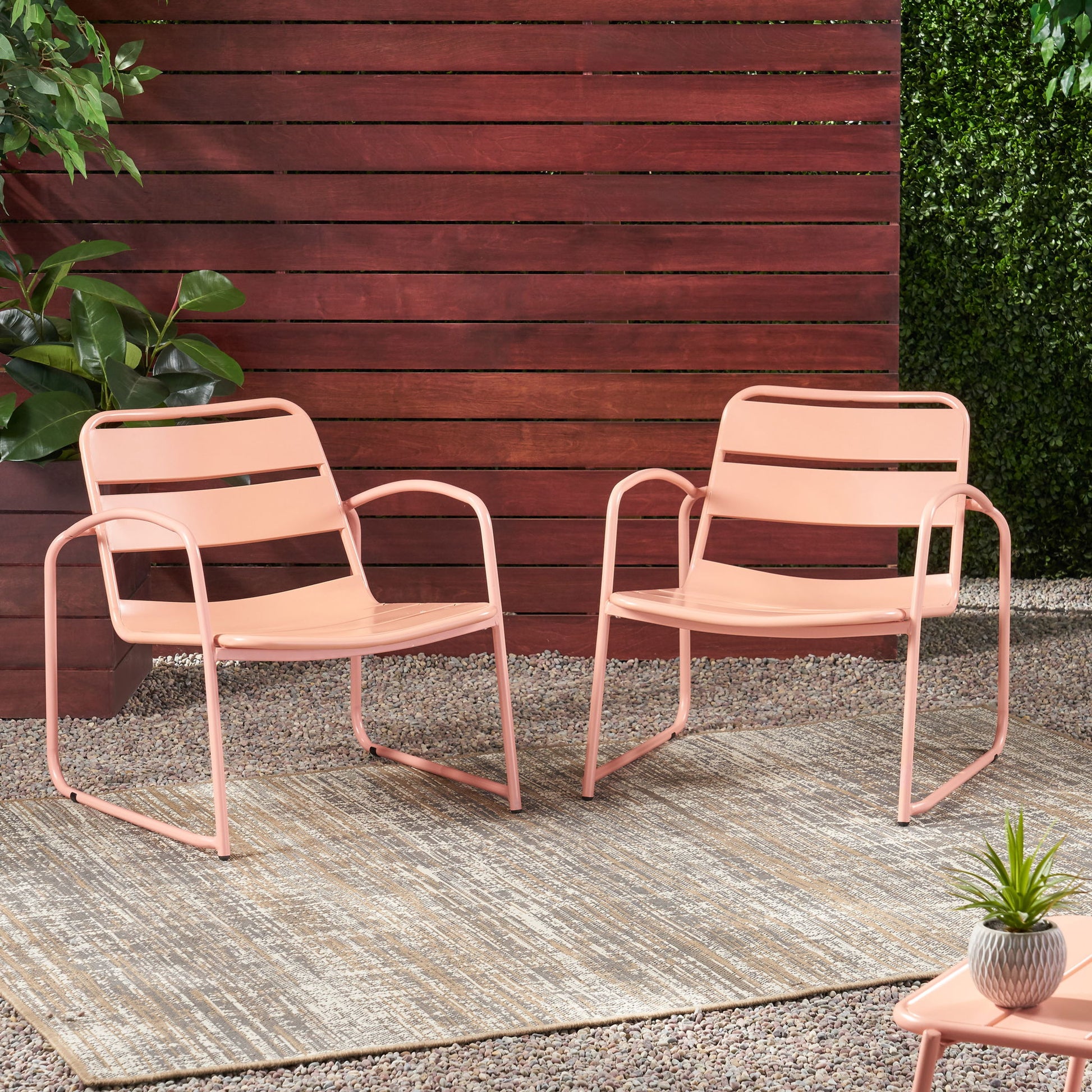 Two Conrad outdoor chairs in pink metal sit on a rug over gravel, near a wooden fence, green plants, and a small table topped with a potted succulent.