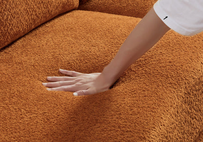 A hand presses down on the soft, textured surface of the Paton - Comfortable Sofa’s orange cushion, leaving a visible indent.