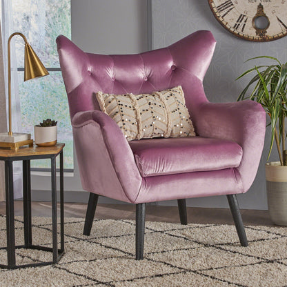The Alyssa Mid Century Upholstered Arm Chair with wingback design in plush pink velvet sits on a geometric rug beside a gold lamp, a small table with a potted plant, and a large wall clock.