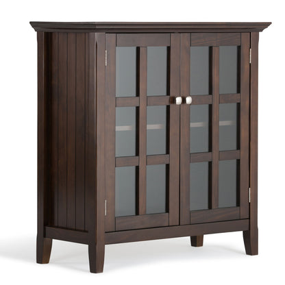 The Acadian Low Storage Cabinet in Brunette Brown features two glass-paneled doors with a grid pattern, silver knobs, square legs, and a modern design. An interior shelf is visible through the doors.