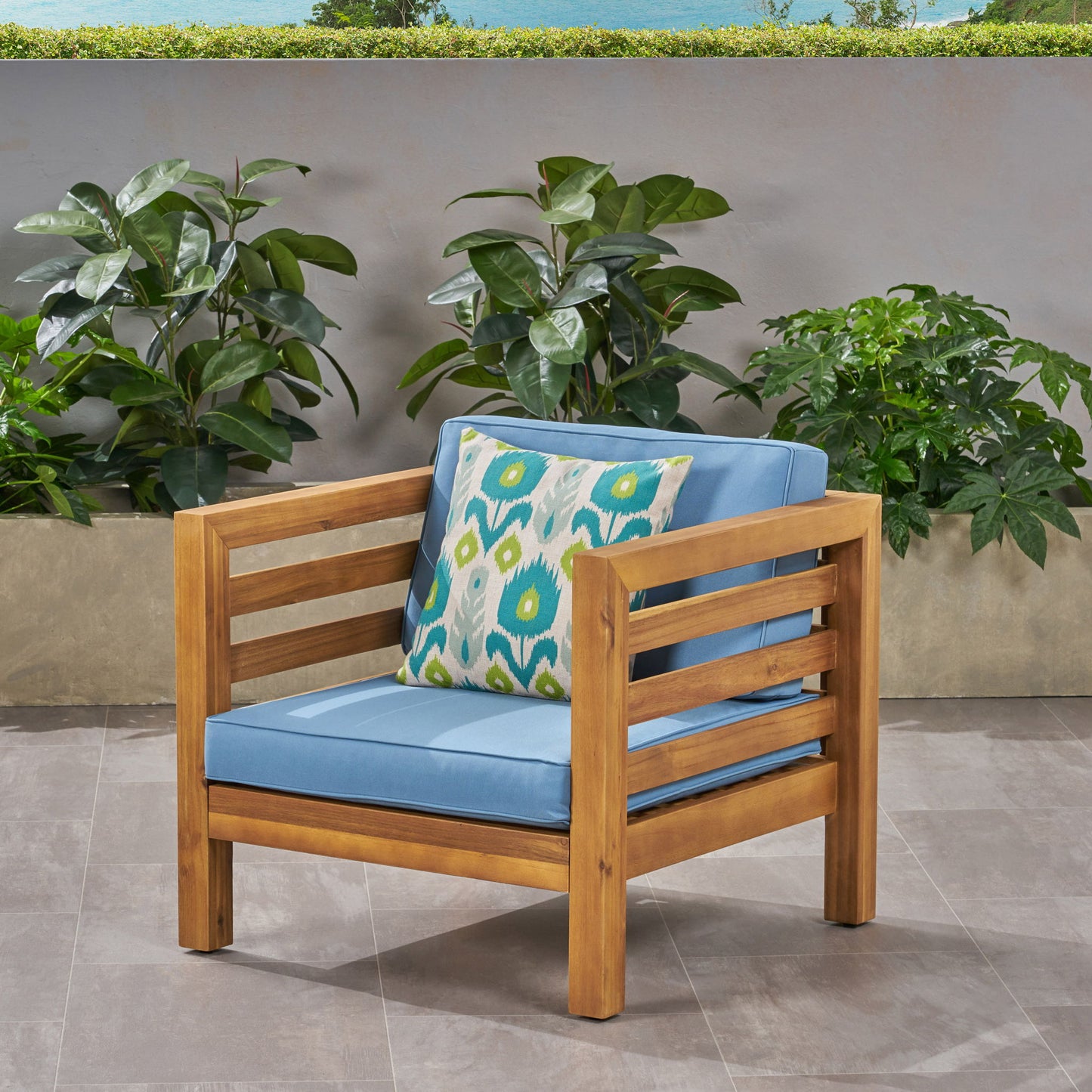 An Outdoor Acacia Wood Patio Chair With Fabric Cushion featuring light blue cushions and a patterned pillow sits on a tiled patio, surrounded by green plants and set against a gray wall.