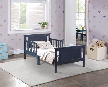 A child’s bedroom featuring the Connelly - Reversible Panel Toddler Bed in navy blue, set on a light gray rug with star-patterned wallpaper, a window with open shutters, a stuffed animal box, and stacked blue suitcases in the corner.