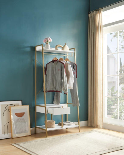 A Metal Clothes Rack Open Wardrobe Free Standing with one drawer and hanging rod stands against a teal wall, holding gray tracksuits. Framed art rests nearby, while shelves display flowers and ceramics. Sunlight streams through a large window.