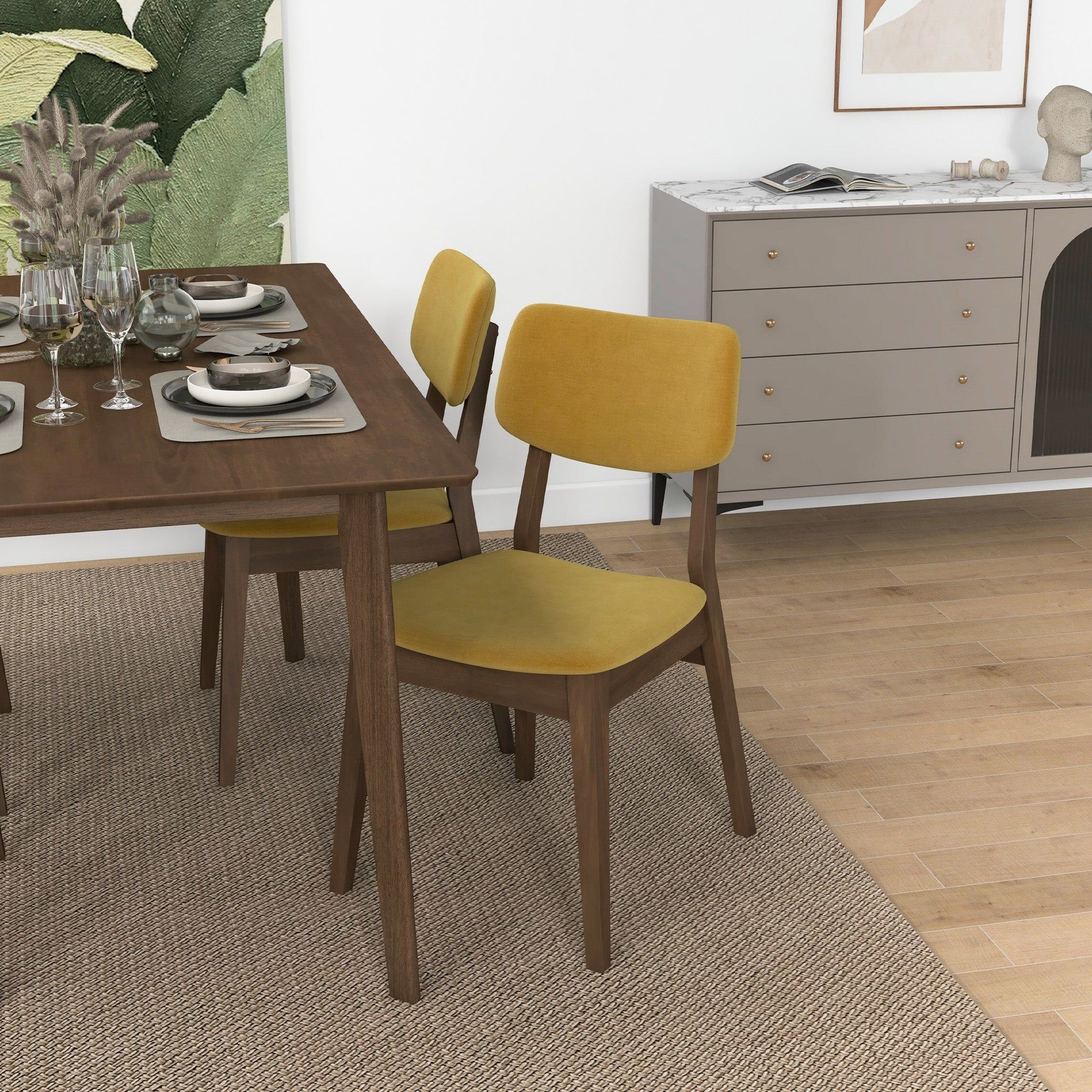 A dining area with a wooden table set for four, featuring yellow Carlos - Solid Back Side Chairs, a beige rug, a modern sideboard, framed art on the white wall, plus plants and tableware on the table.