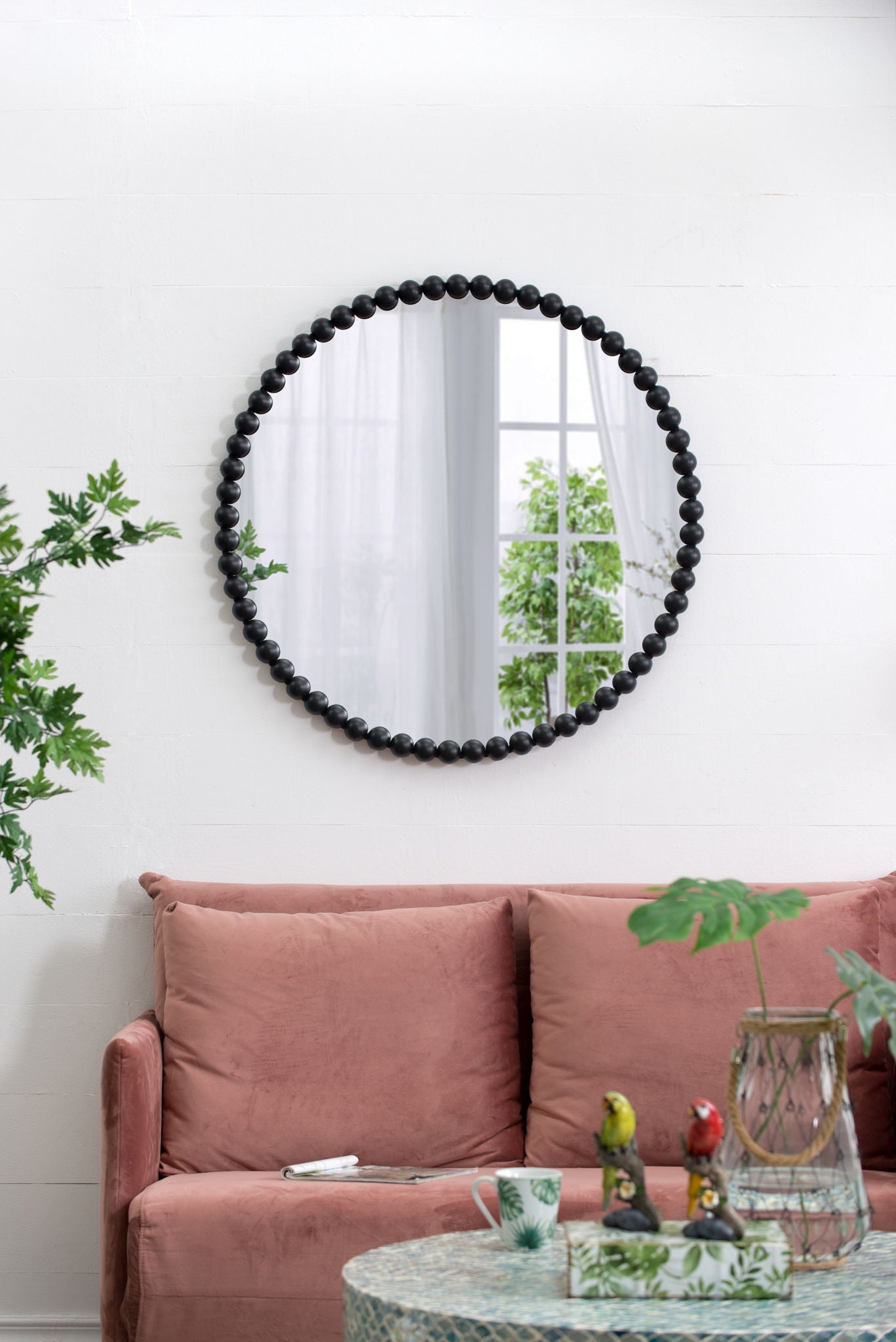 The Circle Mirror With Metal Beaded Frame in black hangs on a white wall above a pink sofa, with a plant to the left and a coffee table in front. The mirror reflects a window and greenery.