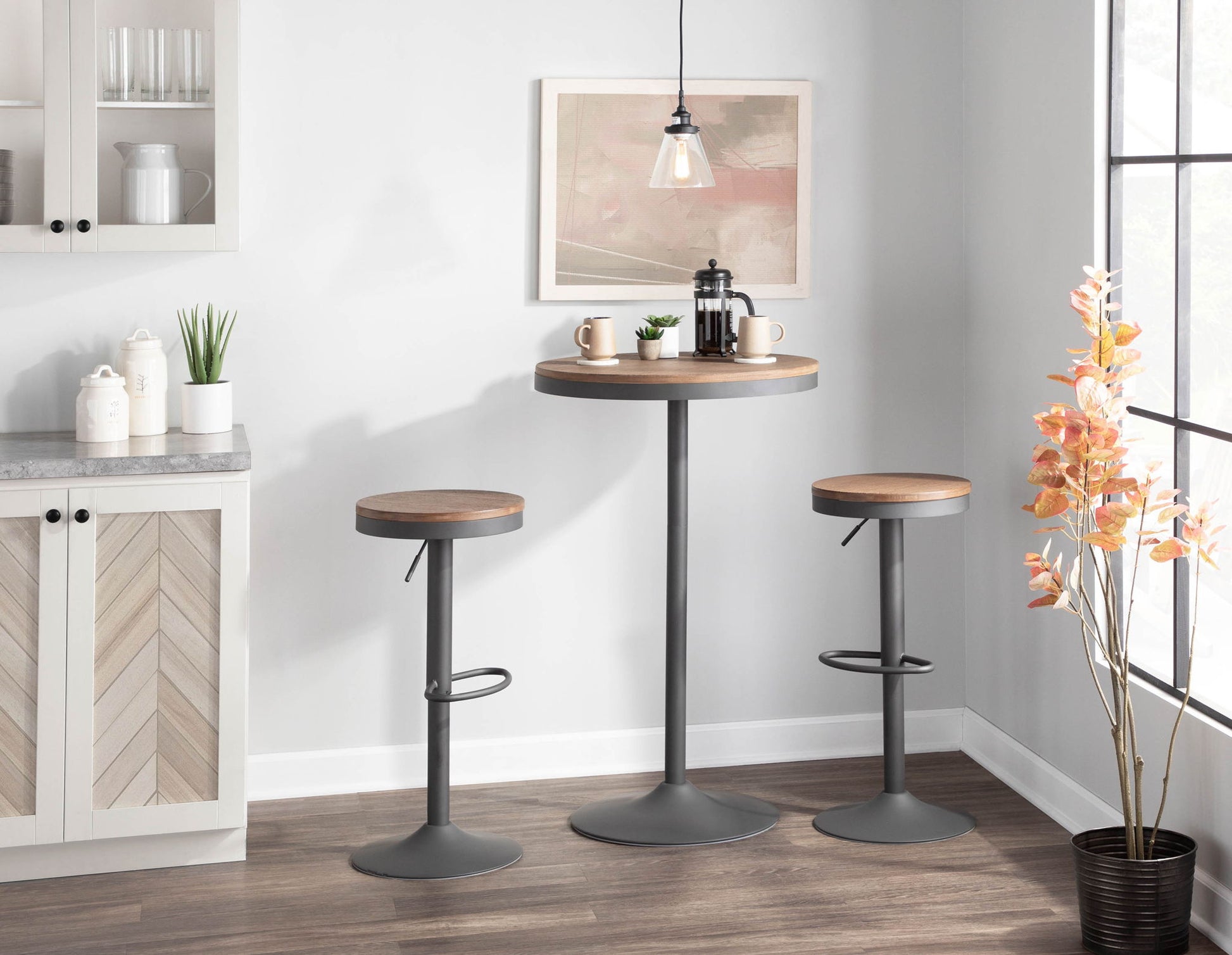 A modern kitchen corner features the Dakota - Industrial Adjustable Bar Dinette Table, paired with two stools, a French press, mugs, a pendant light, wall art, a potted plant, and natural light from a nearby window.