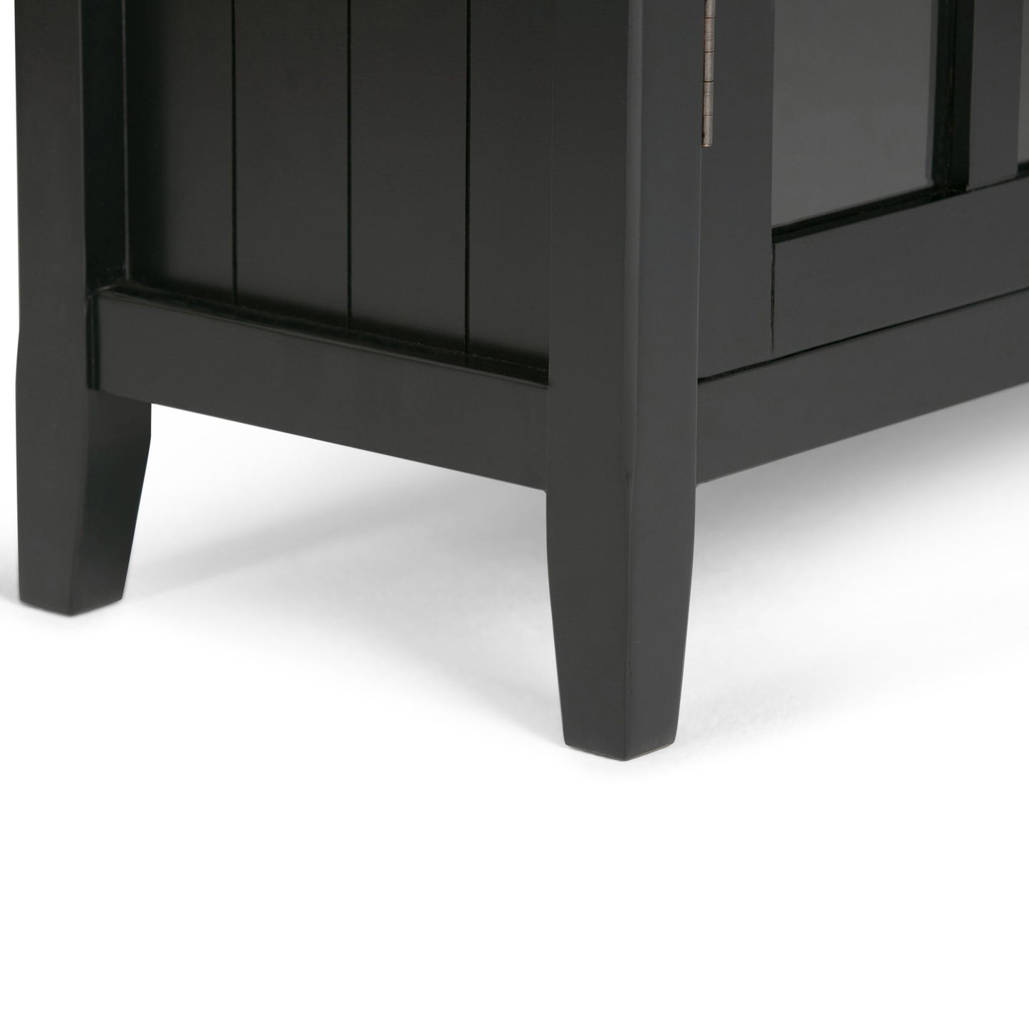 Close-up of the bottom corner of the Acadian - Medium Storage Cabinet, Handcrafted in black wood, showing two legs and part of the side and front panels on a white surface.