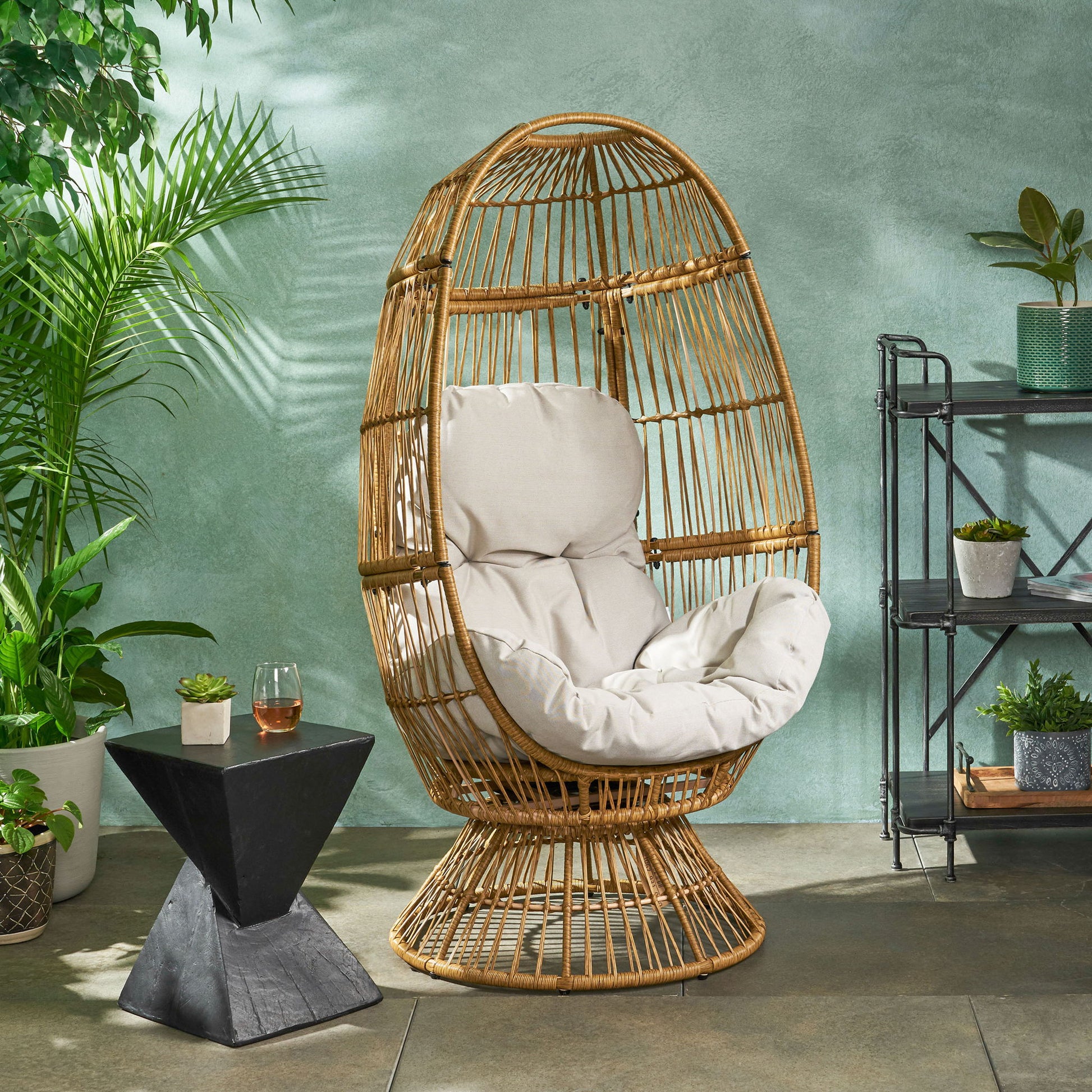 The Patio Swivel Basket Egg Chair With Cushion sits on a patio, surrounded by potted plants and a black geometric side table with a drink, in front of a green wall and black shelving unit filled with more plants.