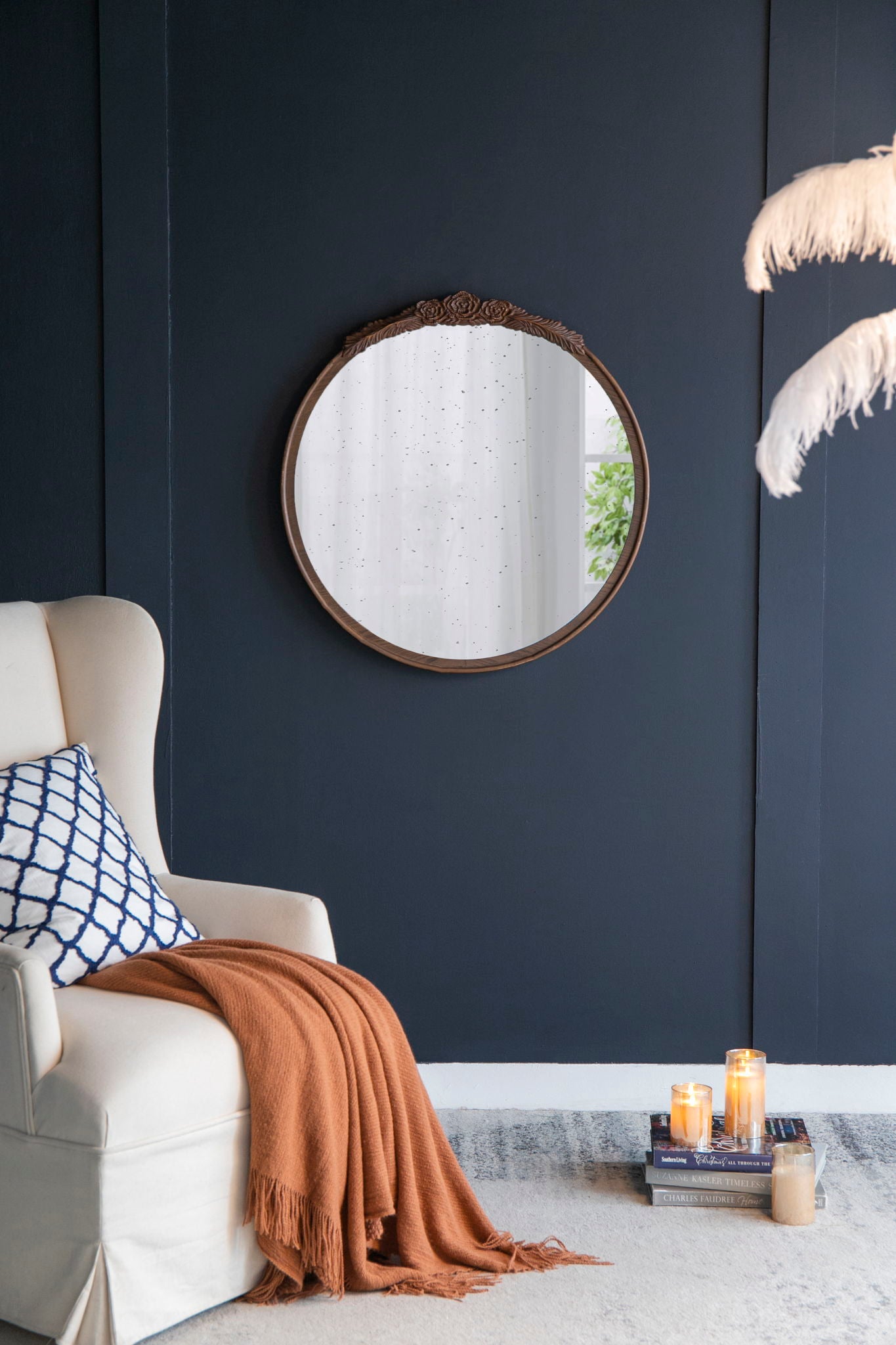 A cozy living room corner features a cream armchair, blue and white cushion, orange throw, and the Round Hand Carved Rose Antique Mirror Frame, Wall Mirror For Live Space hanging above lit candles and stacked books. Feathered decor sits on the right.