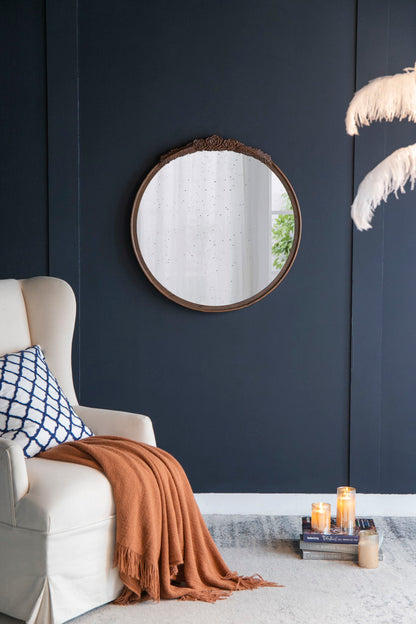 A cozy living room corner features a cream armchair, blue and white cushion, orange throw, and the Round Hand Carved Rose Antique Mirror Frame, Wall Mirror For Live Space hanging above lit candles and stacked books. Feathered decor sits on the right.