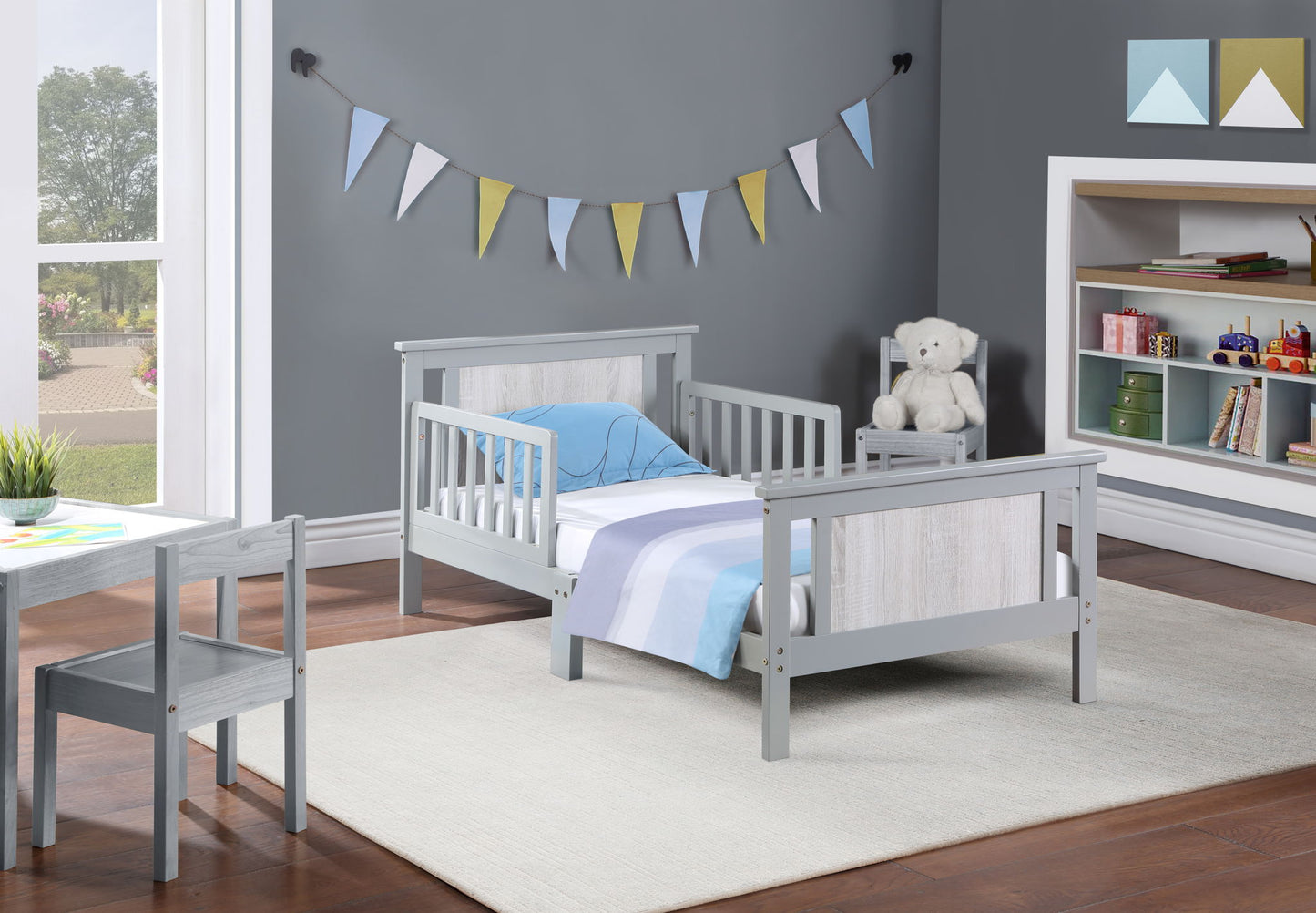A cozy kids’ bedroom features the Connelly Reversible Panel Toddler Bed in gray, striped bedding, a matching chair, shelves with books and toys, a teddy bear on the nightstand, pennant banners on a gray wall, and a white rug.