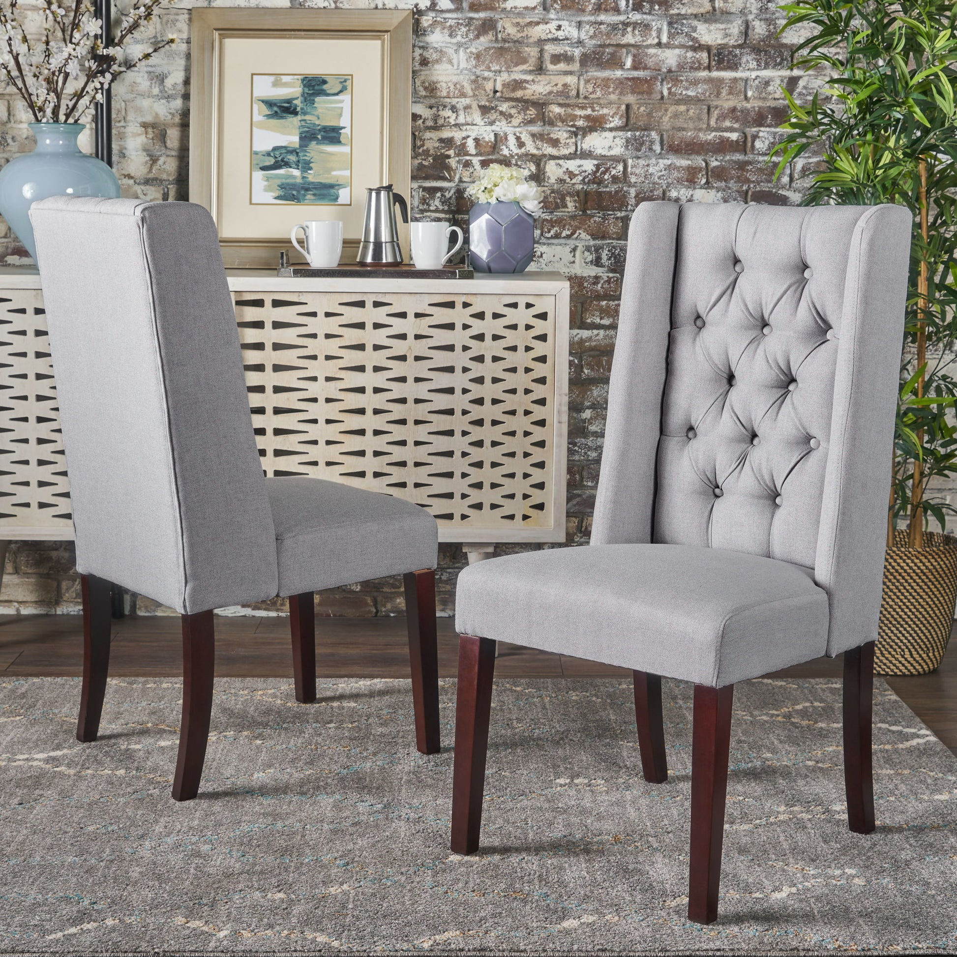 Two Sophisticated Tufted Dining Chairs with high backs and dark wooden legs sit on a patterned rug in a chic room featuring a sideboard, decor accents, and a plant set against an exposed brick wall.