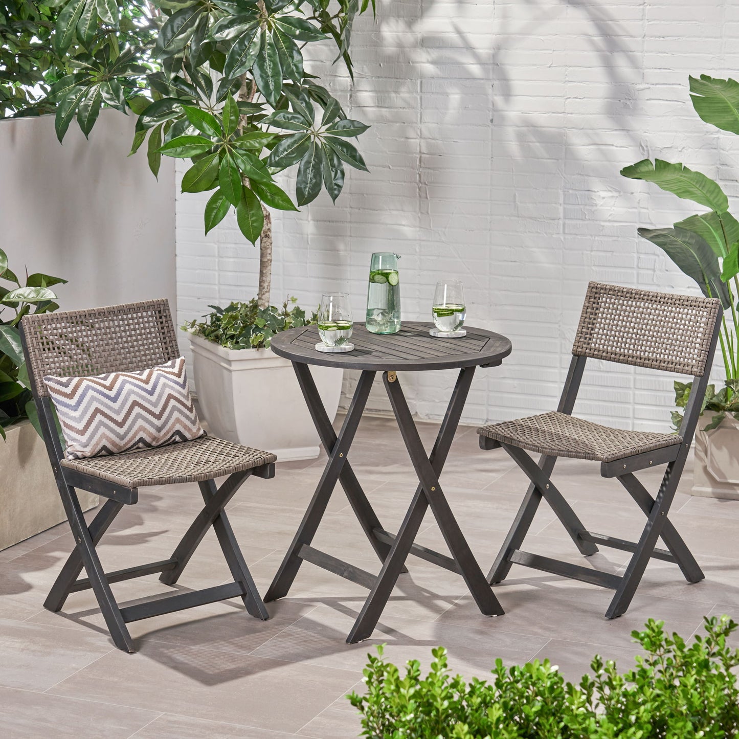 The Evergreen Outdoor Dining Set includes a round dining table and two chairs, styled with pillows, a water carafe, glasses, and surrounded by potted green plants against a white brick wall.