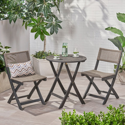 The Evergreen Outdoor Dining Set includes a round dining table and two chairs, styled with pillows, a water carafe, glasses, and surrounded by potted green plants against a white brick wall.