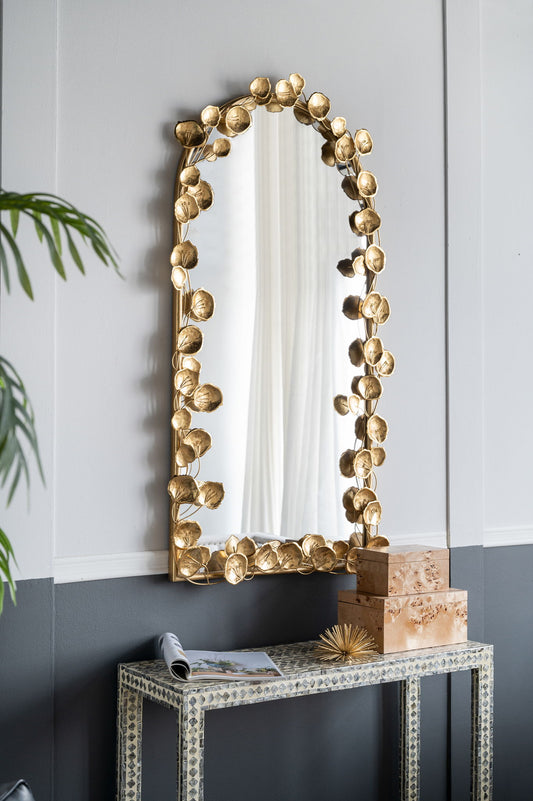 The Full Length Arched Wall Mirror with Leaf Accents in Gold hangs above a console table with a book, two decorative boxes, and a gold spiked ornament in a modern room with gray and white walls.