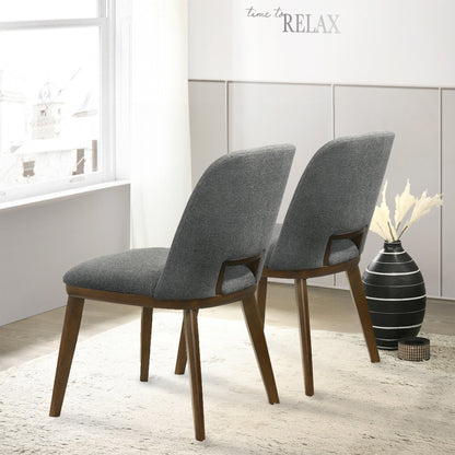 A set of two Blake Modern Dining Chairs with gray upholstery and wooden legs sit on a light rug in a bright room. A black vase with pampas grass is nearby, and "time to RELAX" decorates the wall above.