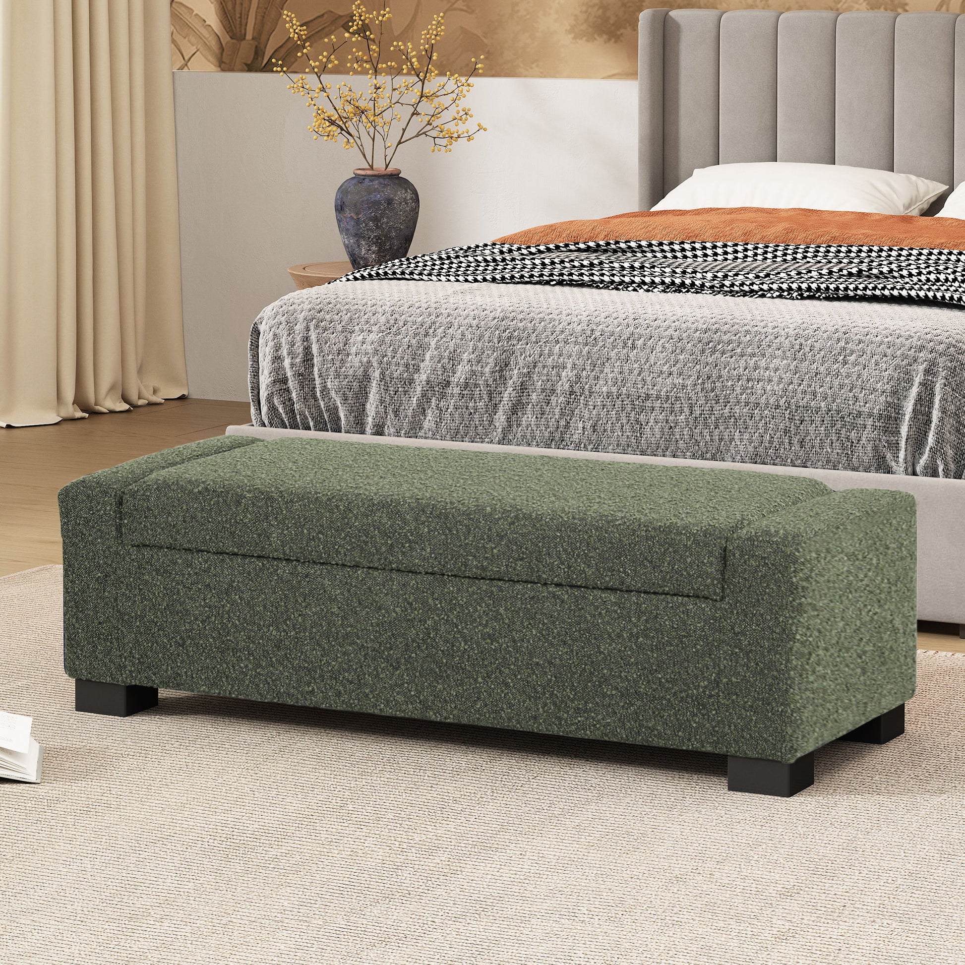 A Textured Fabric Ottoman Bench With Hinged Lid in green sits at the foot of a bed with a gray headboard, beige carpet, and a decorative vase with yellow branches in a modern bedroom.