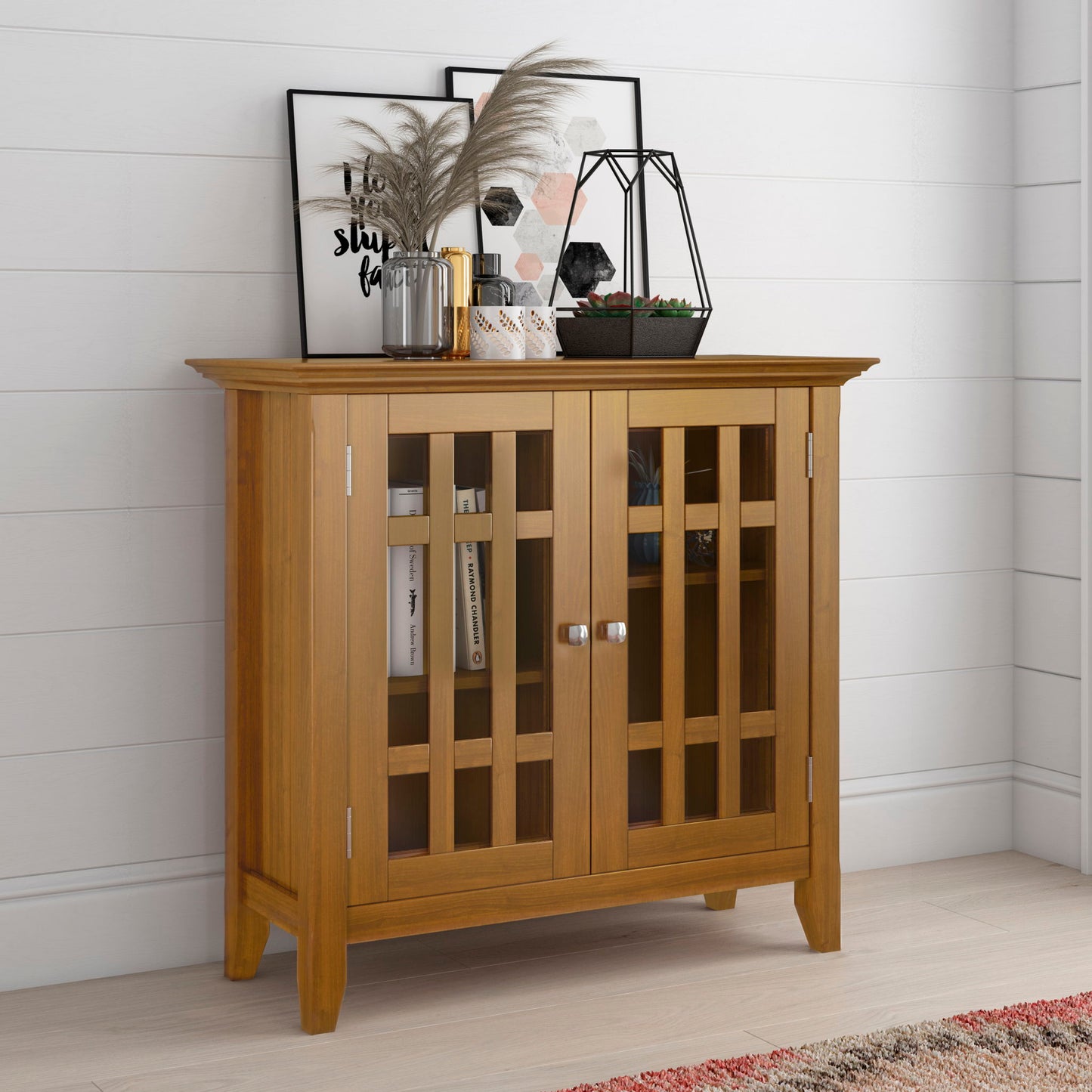 The Bedford Storage Media Cabinet stands against a white wall, topped with framed art, a geometric planter with succulents, a candle, pampas grass in a vase, and other decor. Books are neatly stored inside the glass-paneled cabinet.