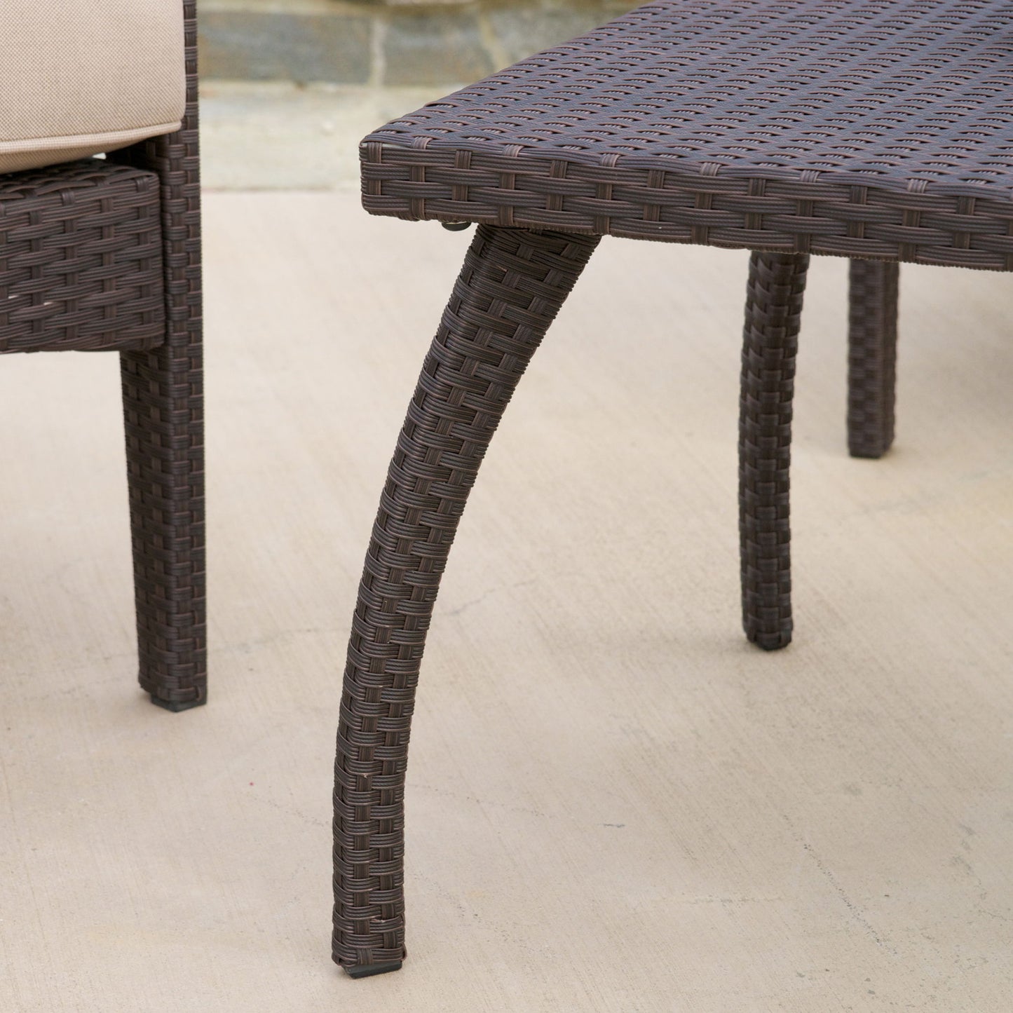 Close-up of the Honolulu - Wicker Set Outdoor Seating, showing its brown woven pattern and curved legs on a light patio. Only parts of the table and chair are visible.