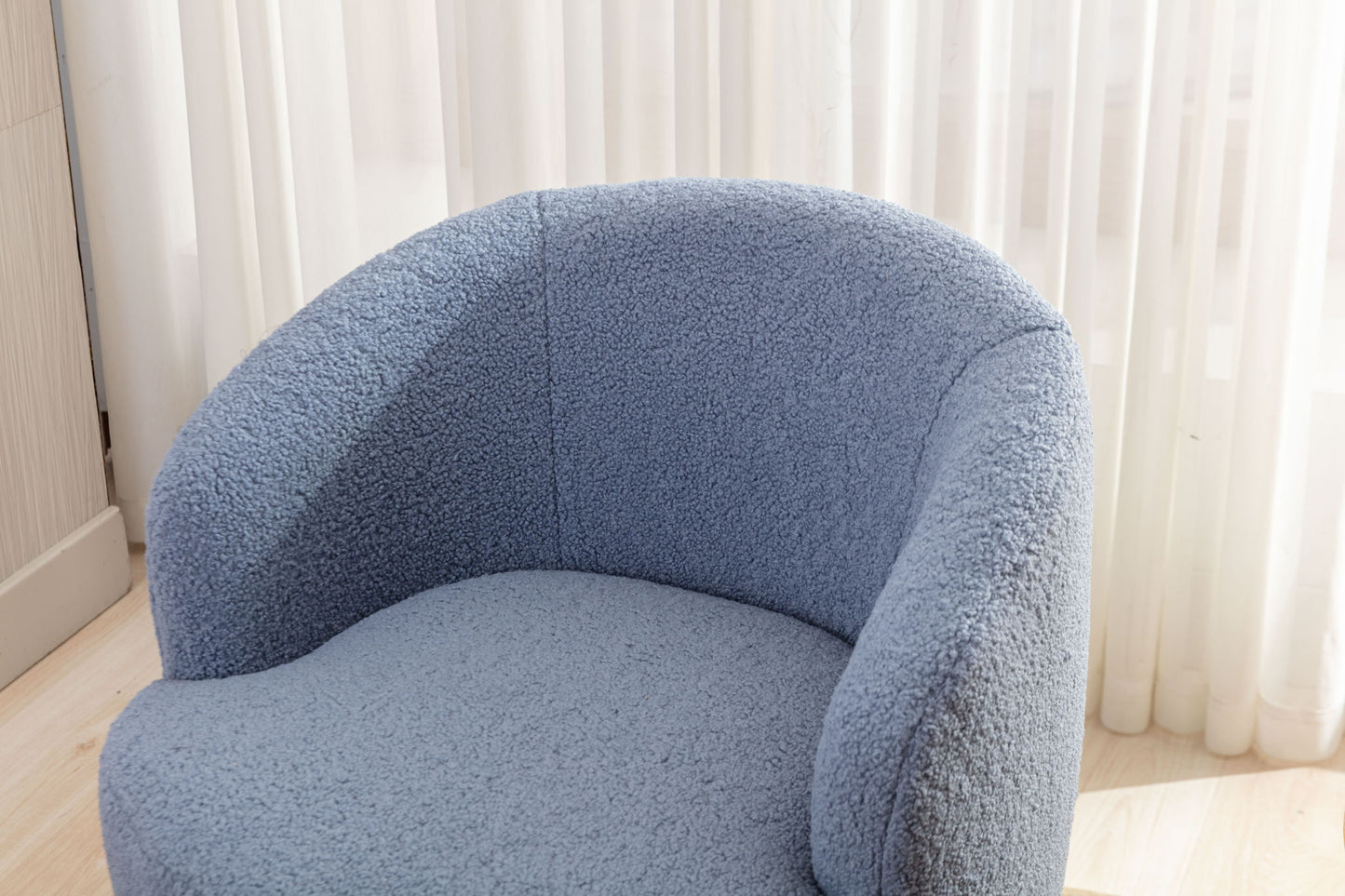 The Teddy Fabric Swivel Accent Armchair Barrel Chair with a powder coating metal ring features textured, light blue fabric and a modern curved design, placed in front of sheer white curtains on light wood flooring in a bright room.