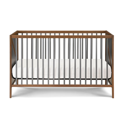 The Pixie Zen 3-in-1 Crib in walnut features a modern wooden frame, vertical dark gray slats, and comes with a white mattress inside.