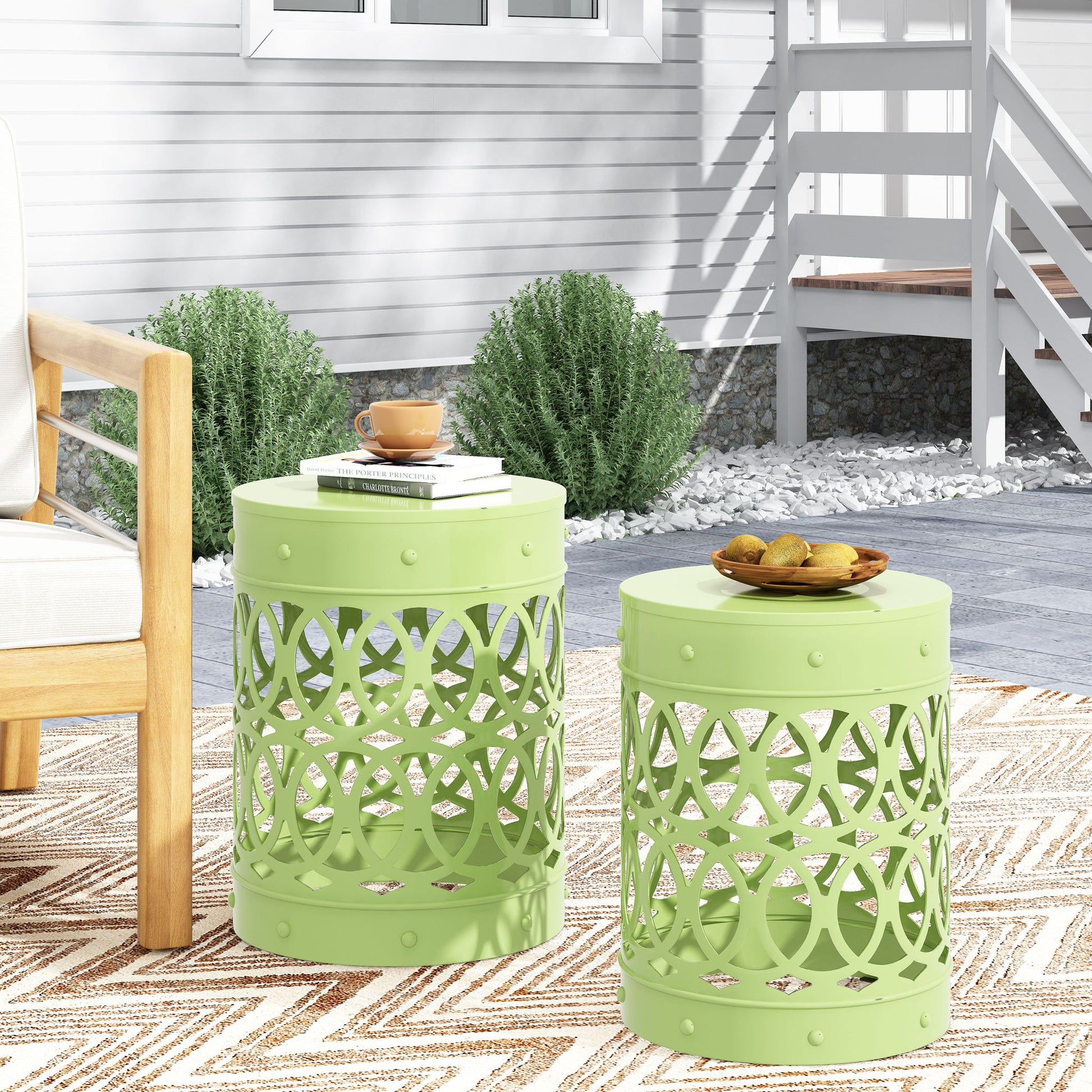 Two light green Outdoor Metal S/2 End Tables with decorative cut-outs stand on a beige geometric rug, one holding a cup, saucer, and book, the other with a plate of snacks. Shrubs and a white house are visible in the background.