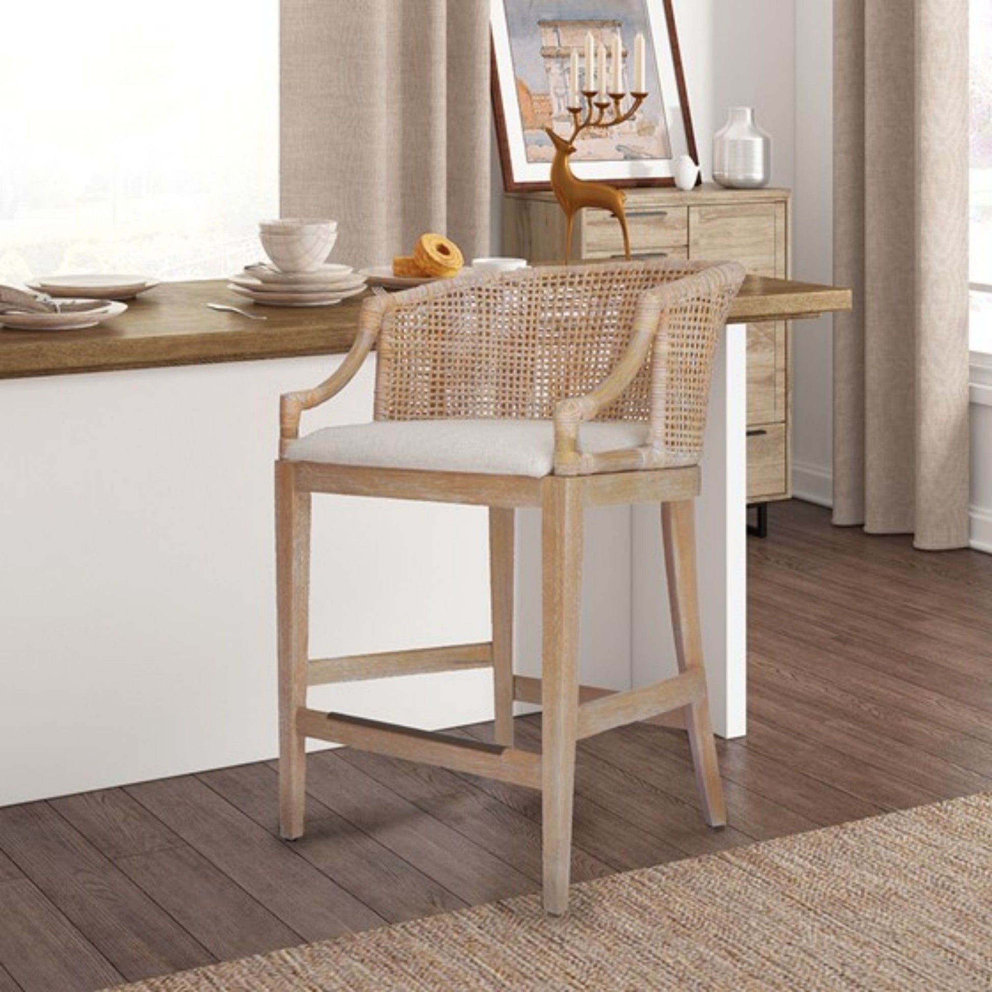 The Casual Handcrafted Rattan Counter Stool, featuring a woven rattan backrest and light cushion, is placed at a kitchen counter, complemented by neutral-toned decor and a sideboard with art and candles in the background.