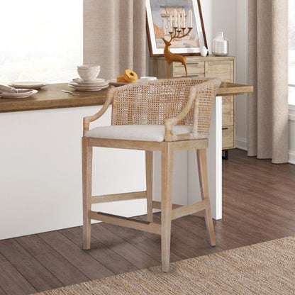The Casual Handcrafted Rattan Counter Stool, featuring a woven rattan backrest and light cushion, is placed at a kitchen counter, complemented by neutral-toned decor and a sideboard with art and candles in the background.