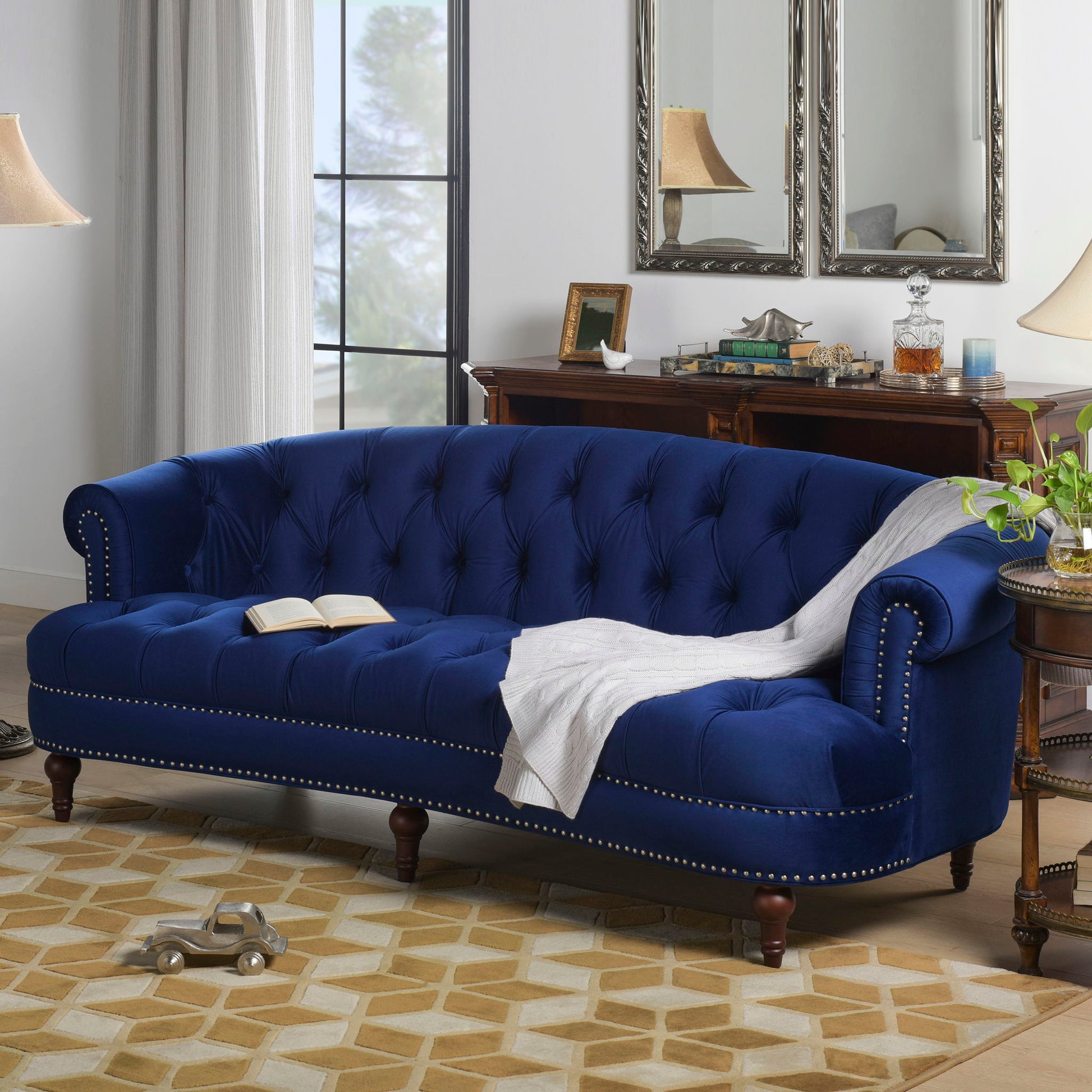 The La Rosa - Victorian Chesterfield Tufted Sofa in blue velvet, featuring studded trim, is styled with a white throw and open book. The cozy living room includes a geometric rug, wooden furniture, lamps, mirrors, and a window.
