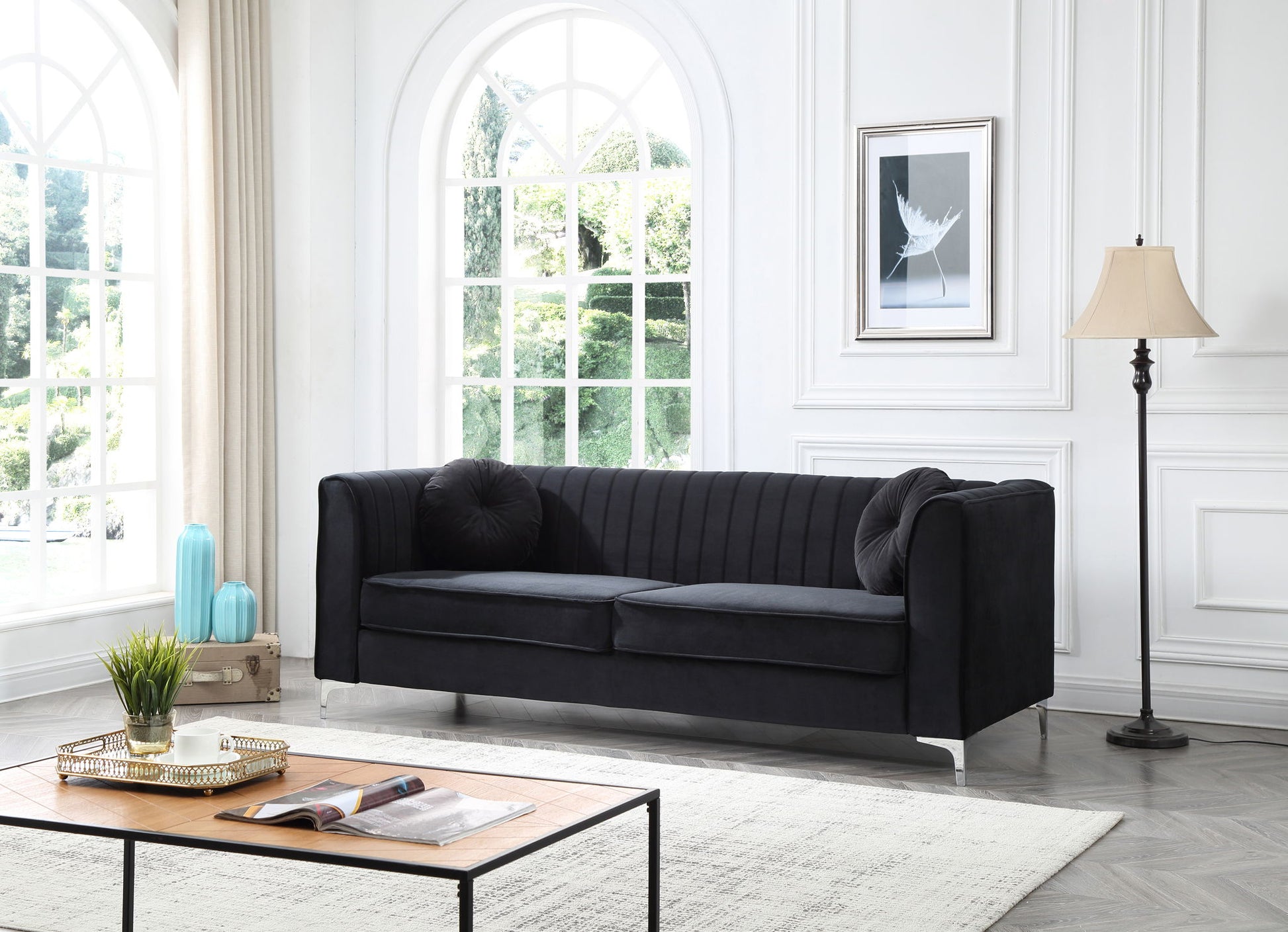 A modern living room with the Delray - Micro Suede Sofa in black, two matching cushions, a wooden coffee table with books and a plant, light walls, large arched windows, framed art, blue vases, and a floor lamp.