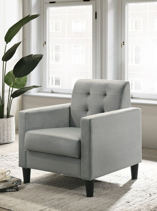 The Hale Velvet Accent Armchair with tufted backrest in light gray is placed on a textured rug by large windows. A potted plant is nearby, and a cup and saucer rests on the rug, adding to the cozy ambiance.