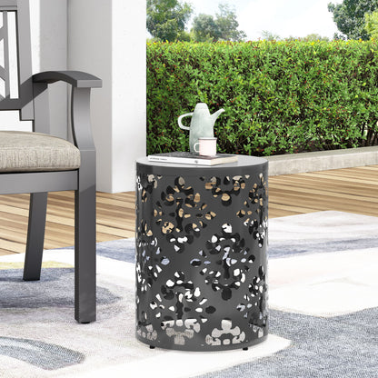 An Outdoor Metal Modern Design End Table with decorative cutouts sits on a patio rug beside a cushioned chair. A teapot and books rest on the table, with green bushes and trees visible in the background.
