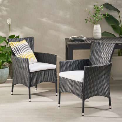 Two Dimensional Outdoor Dining Chairs with cushion seats and iron frames (set of 2) sit on a patio; one features a yellow and gray pillow. A black table with a vase, books, and potted plants stands behind them against a neutral wall.