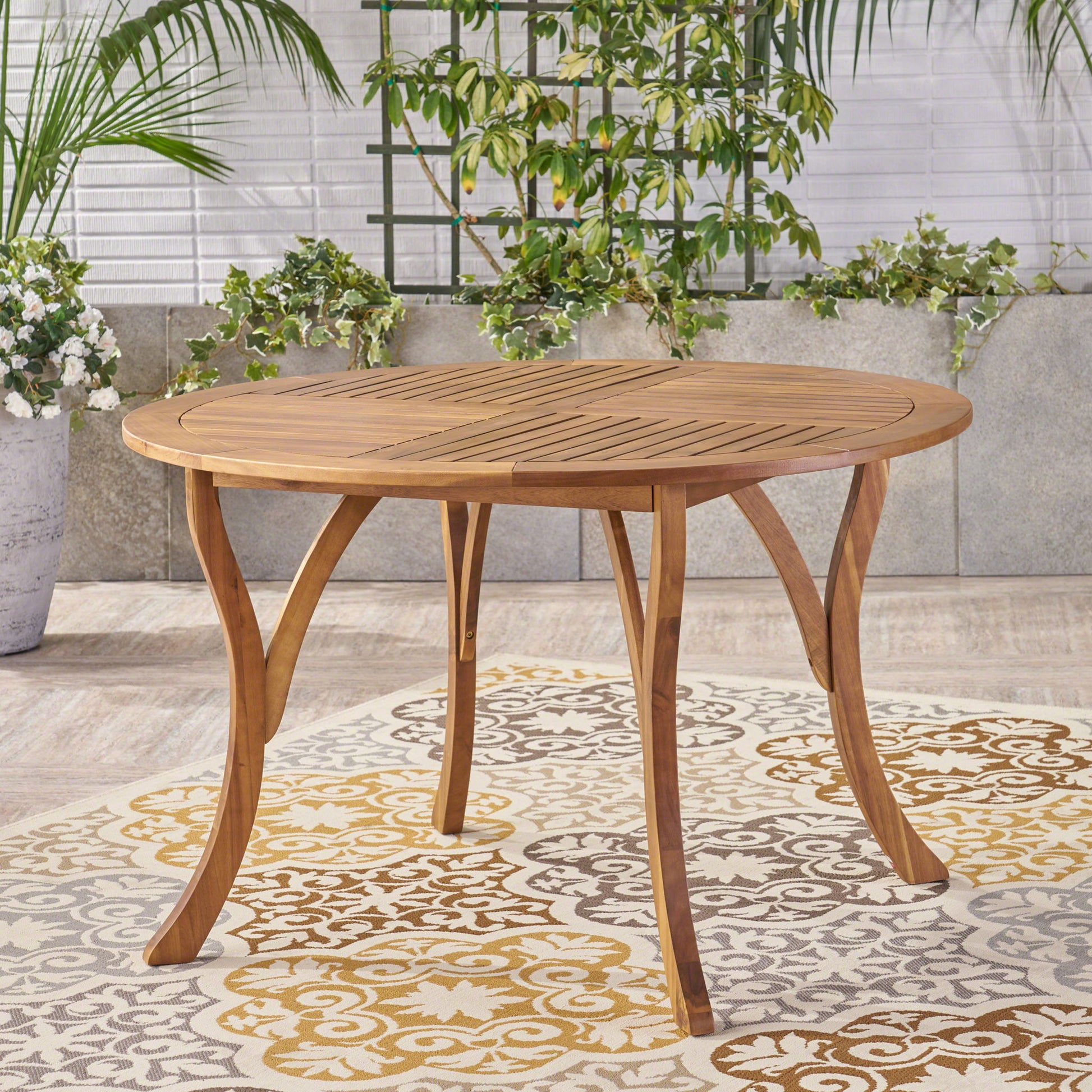 The Hermosa Outdoor Acacia Wood Table features a circular design with curved legs, set on a patterned rug among potted plants and greenery against a stone and white brick wall.