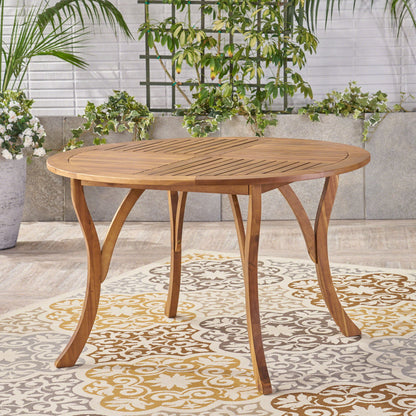 The Hermosa Outdoor Acacia Wood Table features a circular design with curved legs, set on a patterned rug among potted plants and greenery against a stone and white brick wall.