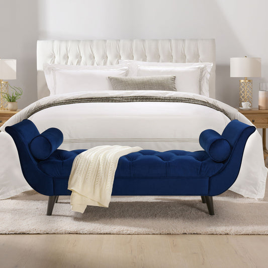 A modern bedroom features a white tufted bed, layered white and gray bedding, wooden nightstands with lamps, and the Alma - Tufted Flared Arm Entryway Bench in blue velvet with a cream knit blanket at the foot of the bed.
