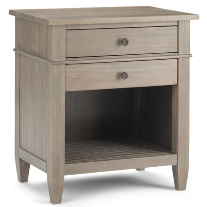The Carlton - Bedside Table, Handcrafted, is a wooden nightstand with a light brown finish, featuring one drawer with a round knob, an open lower shelf, and tapered legs.